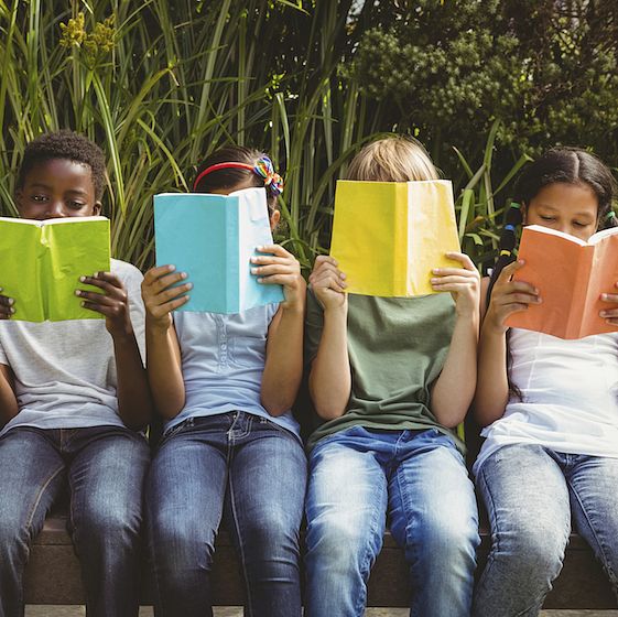 Image of a group of children sitting down and holding books in front of their faces. 