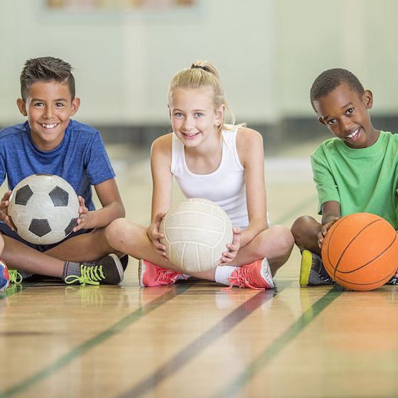 Image of a diverse group of children sitting on a gym floor and holding sports balls. 