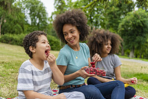 A mother and her two kids laugh while picnicking on cherries outside