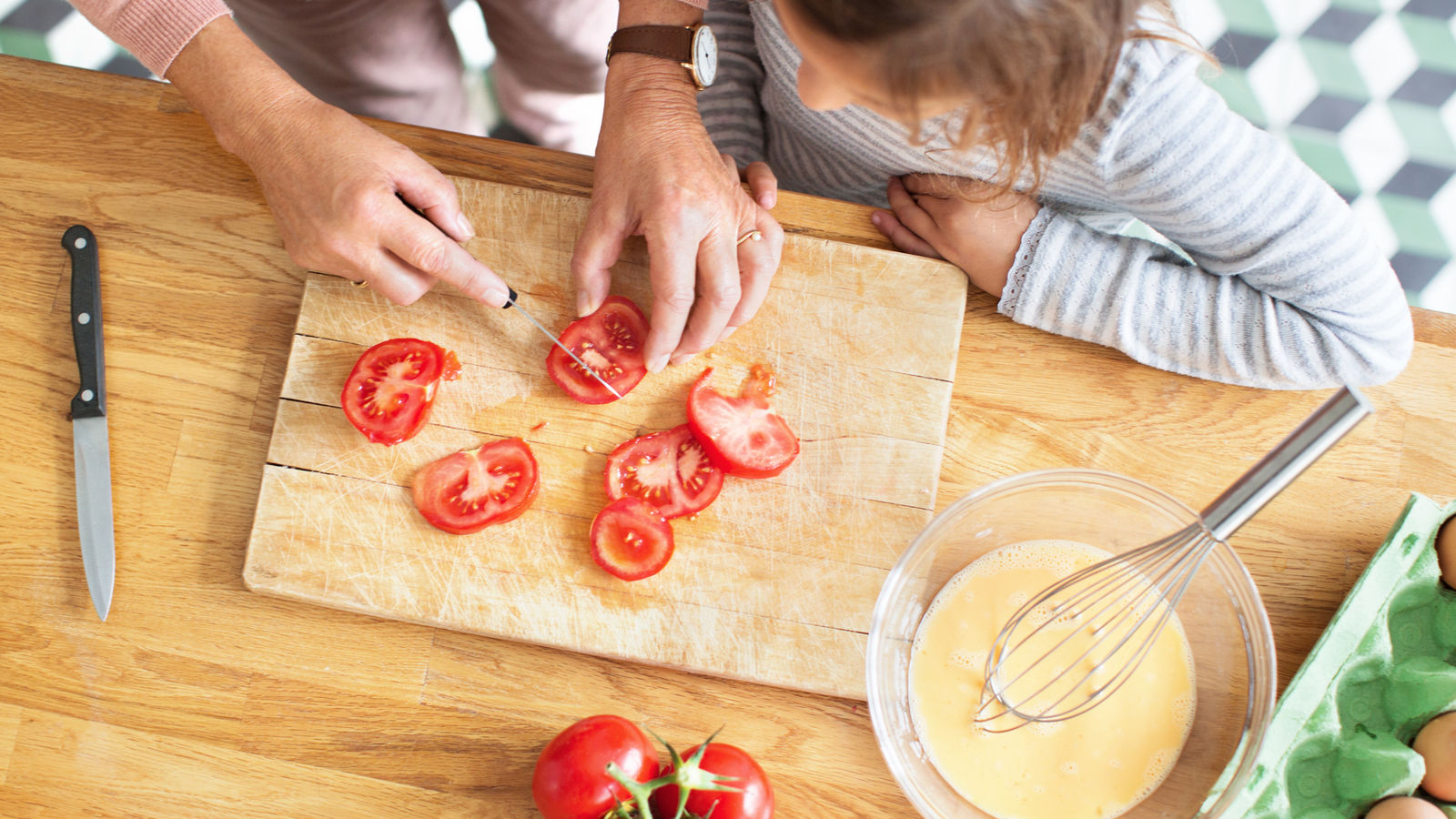 Family cooking with child learning to prepare and cut vegetables.