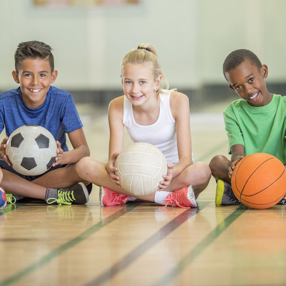 Image of a diverse group of children sitting on a gym floor and holding sports balls. 