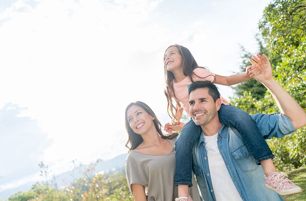 Image of family of three at a park. A young girl rests on top of father's shoulders while the mother stands beside them. 
