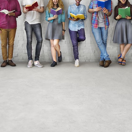 Image of college students leaning against a wall holding books. 